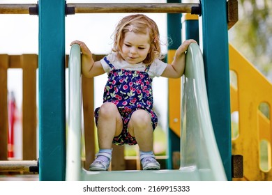 Cute Toddler Girl Playing On Slide On Outdoor Playground. Beautiful Baby In Colorful Shorts Trousers Having Fun On Sunny Warm Summer Day. Child Sliding Down