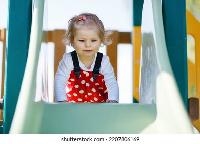 Cute Toddler Girl Playing On Slide On Outdoor Playground. Beautiful Baby In Red Gum Trousers Having Fun On Sunny Warm Summer Day. Child Sliding Down