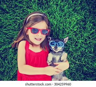 a cute toddler girl with red sunglasses on holding a chihuahua with goggles on in the grass in a park or backyard with a green lawn toned with a retro vintage instagram filter effect app or action 