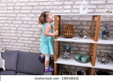 Cute Toddler Girl Standing On Sofa And Reaching For Toys On Shelf