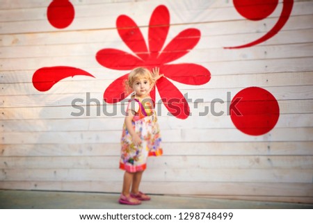 Cute toddler girl stands in front of red and white decorated wooden wall, looking at camera. Colorful summer dress.