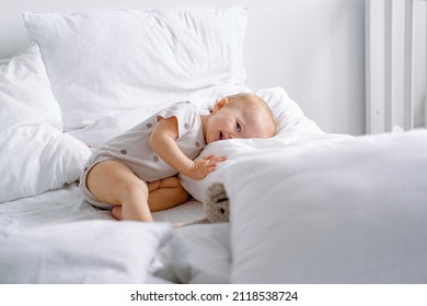 Cute toddler laying on a white bed with lots of pillows and sly smiling looking at camera. 