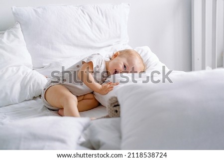 Cute toddler laying on a white bed with lots of pillows and sly smiling looking at camera. 