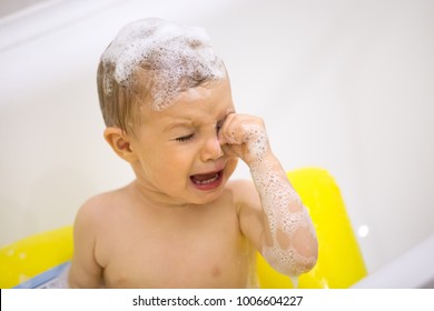 Cute toddler in tears crying taking a bath in bathtub, scared kid afraid of water in bathroom touching irritated by shampoo foam eyes, sad emotional baby screaming weeping in tantrum while bathing