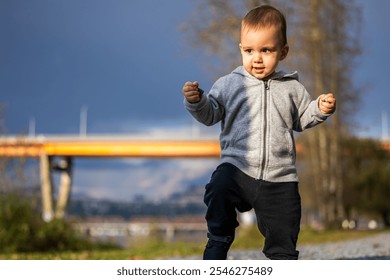 A cute toddler wearing a grey hoodie and dark pants takes confident steps outdoors near a bridge in Mission, BC, Canada. The cloudy sky adds a dynamic backdrop. - Powered by Shutterstock