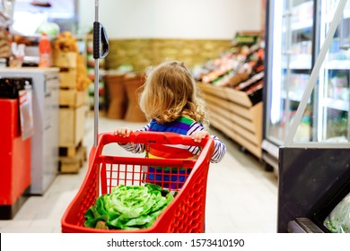 Cute Todler Girl Pushing Shopping Cart In Supermarket. Little Child Buying Fruits. Kid Grocery Shopping. Adorable Baby Kid With Trolley Choosing Fresh Vegetables In Local Store.