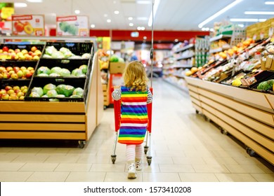 Cute Todler Girl Pushing Shopping Cart In Supermarket. Little Child Buying Fruits. Kid Grocery Shopping. Adorable Baby Kid With Trolley Choosing Fresh Vegetables In Local Store.