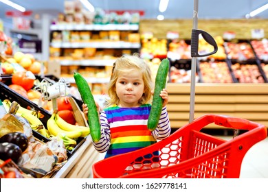 Cute Todler Girl Pushing Shopping Cart In Supermarket. Little Child Buying Fruits. Kid Grocery Shopping. Adorable Baby Kid With Trolley Choosing Fresh Vegetables In Local Store.