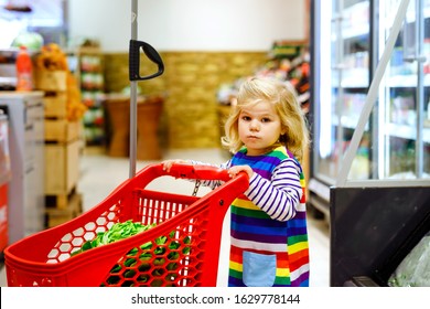 Cute Todler Girl Pushing Shopping Cart In Supermarket. Little Child Buying Fruits. Kid Grocery Shopping. Adorable Baby Kid With Trolley Choosing Fresh Vegetables In Local Store.