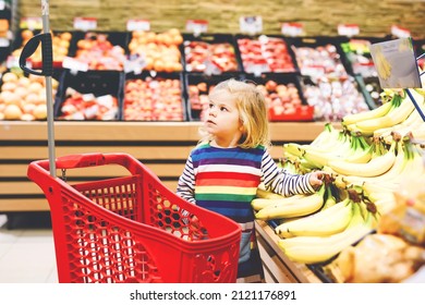 Cute Todler Girl Pushing Shopping Cart In Supermarket. Little Child Buying Fruits. Kid Grocery Shopping. Adorable Baby Kid With Trolley Choosing Fresh Vegetables In Local Store.