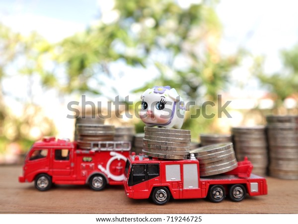 Cute toy piggy on rolls of\
coins stands on miniature fire truck car and blur step ladder of\
money on wood table in natural garden tree and bright light in blue\
sky 