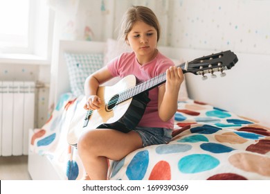 Cute tween girl in pink t-shirt play guitar sit on bed in bright room at home
