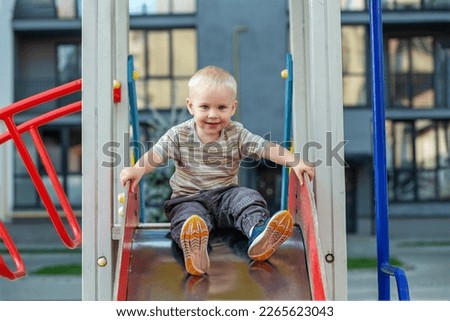 Cute two year old boy plays on playground slide on summertime. Kid play on kindergarten yard. Active kid on colorful slide and swing.