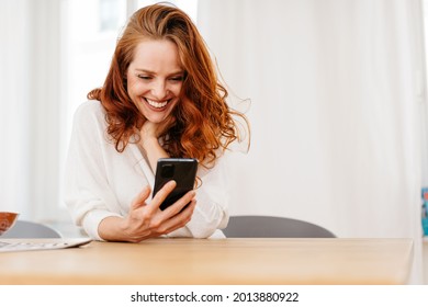 Cute vivacious redhead woman grinning at her smartphone with a beaming smile of pleasure as she reads a text message or watches media seated at a table indoors - Powered by Shutterstock