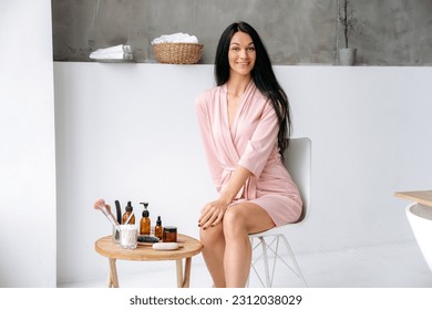 Cute well-groomed caucasian brunette woman in a bathrobe sits in the bathroom near a small table, on the table there are products for skin care, body, face, hair, female posing, smile, looks at camera - Shutterstock ID 2312038029