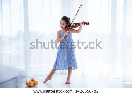 A cute white Asian girl wearing a blue skirt dress is playing a violin in a white background with sunlight.