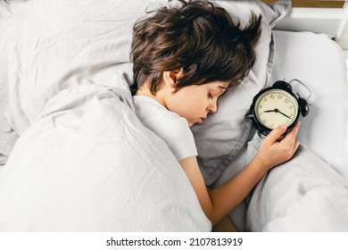 Cute white boy peacefully lying in the bed sleeping early morning after turning off his alarm clock and overslept the school. Early wake up, not getting enough sleep time concept. Top view