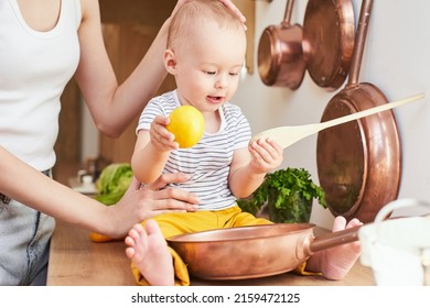 Cute White Son 1 Year Old, Sits On The Table, Plays With His Mother A Frying Pan And A Lemon. A Series Of Photos From Everyday Life In A Real Interior.