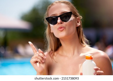 Cute woman with duckface applies sunscreen solar cream on face near swimming pool. Smiling pretty girl puts suntan cream from plastic bottle on skin in spa. Female in bikini with suntan lotion.