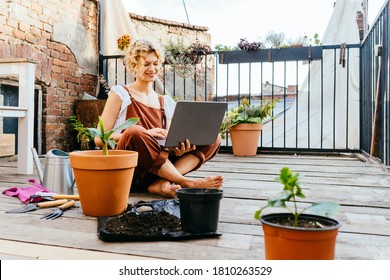 Cute woman gardener wear brown overalls, sitting on wooden floor in terrace resting, using laptop after work, smiling and speaking on video call surrounded by plants. Home gardening concept. - Powered by Shutterstock