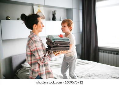 Cute Woman Mom And Son Child With A Stack Of Clean Towels On Hand Against The Background Of A Gray Bedroom. The Concept Of Cleaning, Clean Linen And Waiting For Guests, Help The Child In Housework.