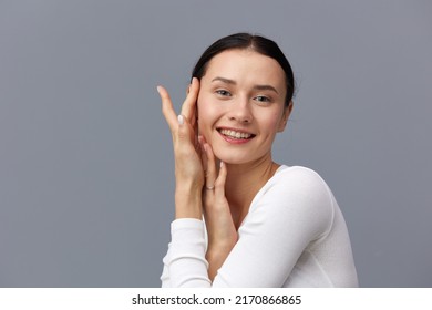A Cute Woman With A Ponytail On Her Head Stands On A Dark Background In A White Tight T-shirt, Put Two Hands On Her Cheek And Smiles Happily