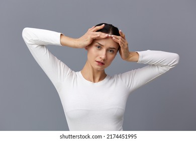 A Cute Woman With A Ponytail On Her Head Stands On A Dark Background In A White Tight T-shirt, Put Her Hands On Her Head And Smiles Happily