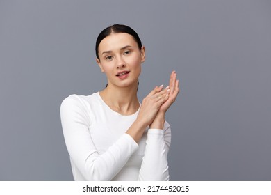 A Cute Woman With A Ponytail On Her Head Stands On A Dark Background In A White Tight T-shirt, Holding Two Hands Together