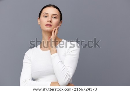 a cute woman with a ponytail on her head stands on a dark background in a white tight T-shirt, put her hand on her neck holding it with the other