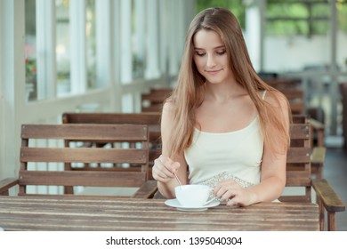 Cute Woman Sitting At The Balcony Terrace Of A Fancy Cafe, Coffee Shop Restaurant Mixing Sugar In Her Cup Of Hot Beverage Tea Or Coffee. Cool Young Modern Female Model.