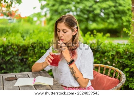 Cute  woman sitting on a chair in summer garden, drinking ice tea and writing in a notebook