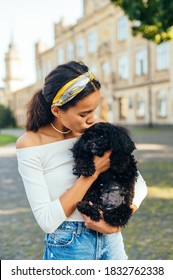 Cute Woman Stands On The Street And Kisses The Dog She Is Holding. Close Portrait Of A Hispanic Woman Holding A Dog And Hugging. Vertical.