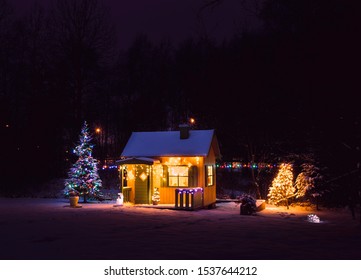 Cute, Wooden Painted Yellow Private Children`s Play House In Home Garden, Decorated With Christmas LED String Lights Outdoors In Cold Snowy Winder Night. Decorated Christmas Fir Tree.