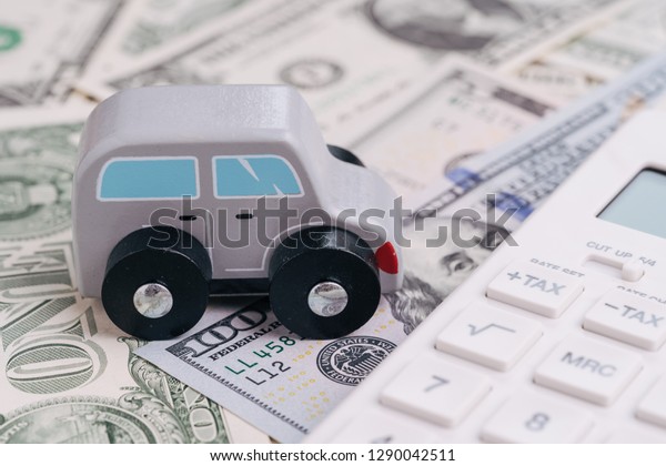 Cute wooden small toy car parking beside white\
calculator on pile of dollar banknotes money, car leasing, rental\
or insurance and maintenance calculation concept, down payment,\
installment for auto.