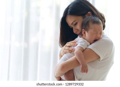Cute yawning Asian infant baby on mothers hands standing in the room near the window. Mother hugging baby with love. Happy motherhood and maternity concept.