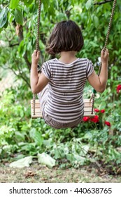 cute young 5-year old kid swinging to relax and play in a backyard for preschooler wellbeing or insouciance in the summertime, back view with tree background
