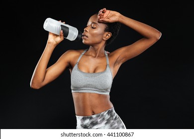 Cute young african american sportswoman standing and holding bottle of water over black background