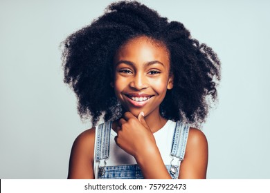 Cute young African girl with long curly hair smiling and wearing dungarees standing with her hand on her chin against a gray background