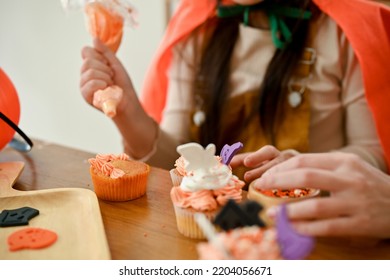 A cute young Asian girl enjoys making a Halloween cupcake with her mom, decorating her cupcake with cream. cropped shot - Powered by Shutterstock