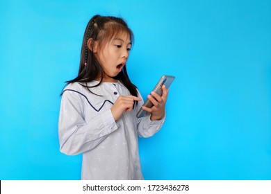 A Cute Young Asian Girl Holding Her Smartphone, Playing And Reading News, Feeling Happy. Plain Light Blue Background. 