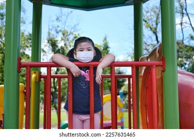 A cute young Asian girl is playing in a jungle gym in a playground during school recess, resting her arms on a metal rail, wearing a face mask for protection against Covid-19. School life in pandemic - Powered by Shutterstock