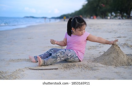 A Cute Young Asian Girl Sitting And Playing Alone On A Sandy Beach, Having Fun, Building A Sand Castle.