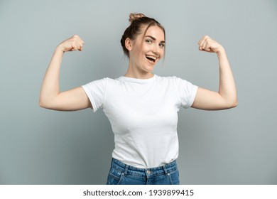 Cute young blonde woman in white t-shirt, blue jeans with on gray background. Strong girl shows muscles, bent arm at the elbow.