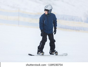 Cute Young Boy, Brave Kid In Gray Helmet And Orange Googles, In Blue Jacket Snowboarding On White Snow Mountain. Winter Sport, Active Lifestyle Concept