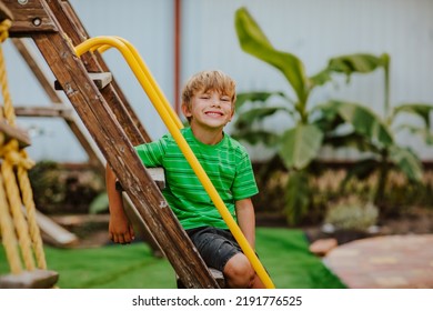 Cute Young Boy In Green Shirt And Black Shorts Is Playing On Back Yard Playground. Copy Space