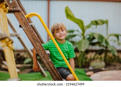 Cute Young Boy In Green Shirt And Black Shorts Is Playing On Back Yard Playground. Copy Space