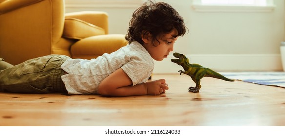 Cute Young Boy Looking At His Dinosaur Toy At Home. Adorable Little Boy Playing With A T-rex Dinosaur Toy While Lying On The Floor In His Play Area.