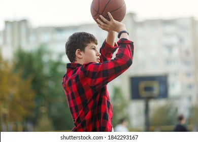 Cute Young Boy Plays Basketball On Street Playground. Teenager In Red Check Flannel Shirt With Orange Basketball Ball Outside. Hobby, Active Lifestyle, Sport Activity For Kids.