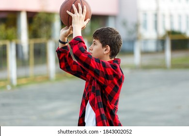 Cute Young Boy Plays Basketball On Street Playground. Teenager In Red Check Flannel Shirt With Orange Basketball Ball Outside. Hobby, Active Lifestyle, Sport Activity For Kids.