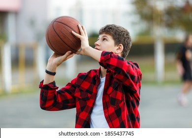 Cute Young Boy Plays Basketball On Street Playground. Teenager In Red Check Flannel Shirt With Orange Basketball Ball Outside. Hobby, Active Lifestyle, Sport Activity For Kids.